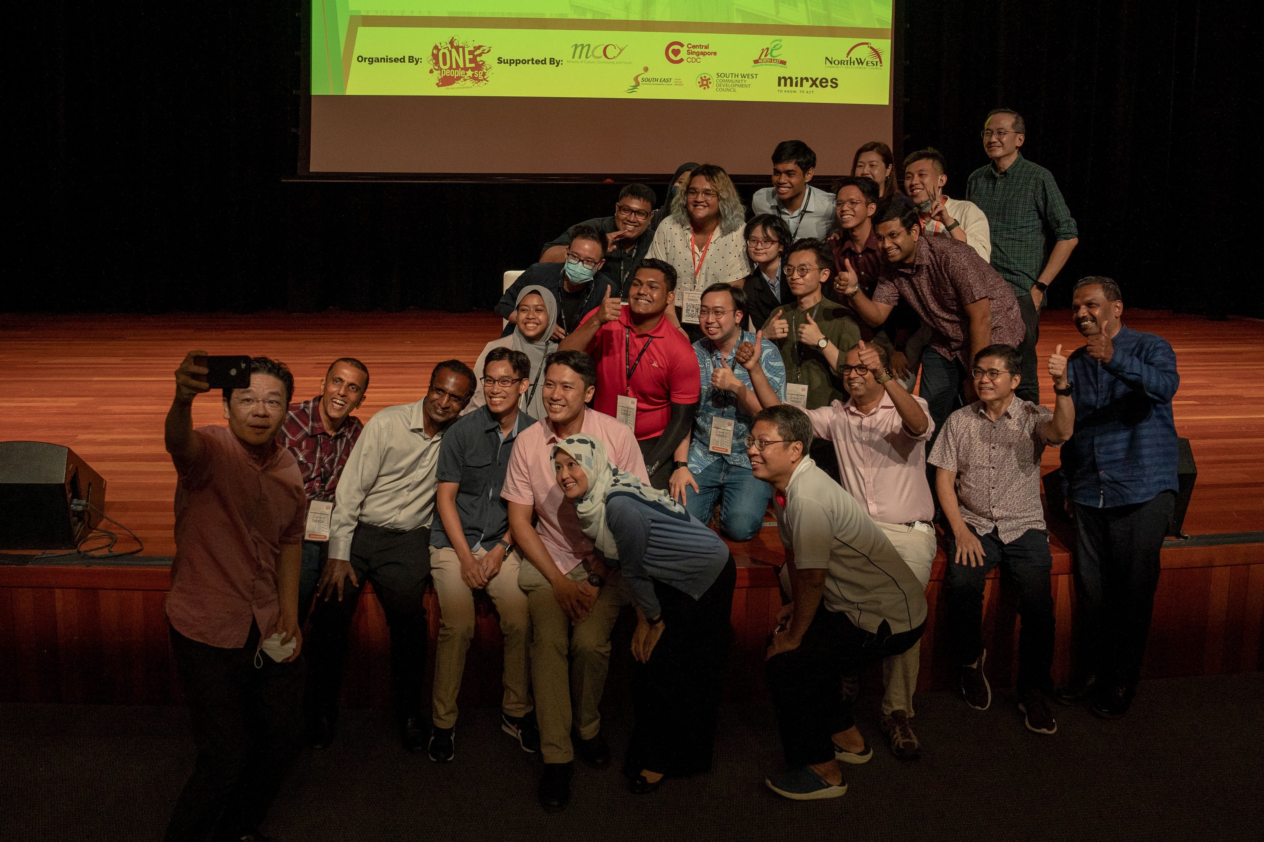 A group of approximately 20 people pose for a photo on a wooden stage with event organizers' logos displayed above.
